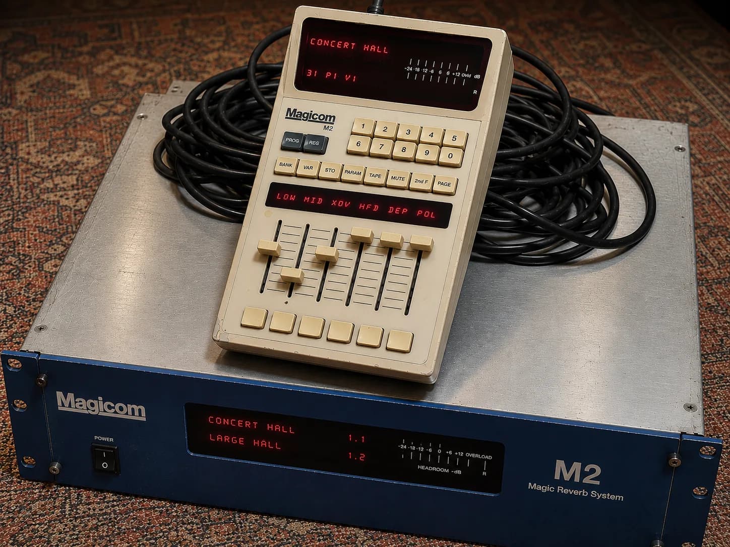 Ilustração da unidade fictícia MAGICOM M2: caixa de reverb de mesa com teclado numérico, display vermelho e faders, cercada por cabos.
